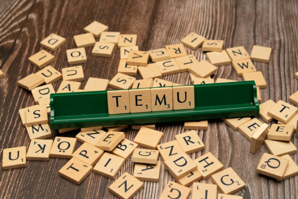 Wooden Scrabble tiles spelling TEMU on a green rack with scattered tiles on a wooden surface.