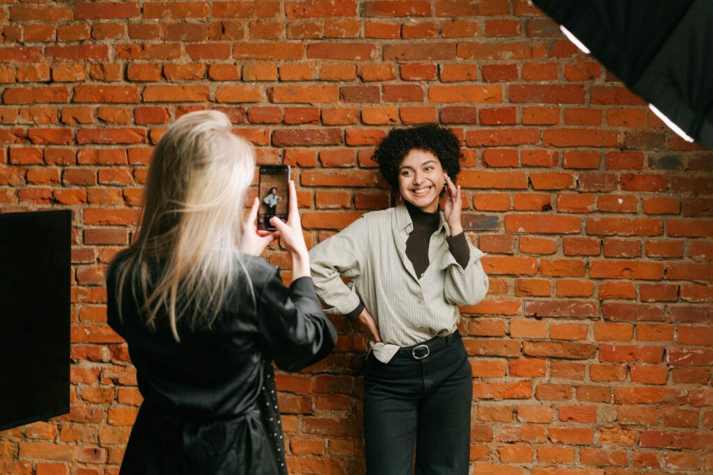 A woman smiles while posing against a brick wall during a photo shoot. Captured indoors.